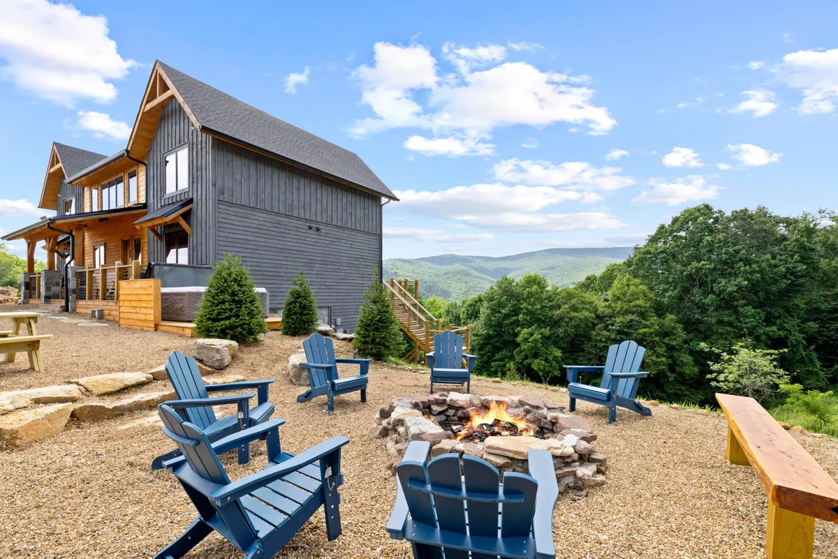 An outdoor fire pit area with Adirondack chairs sits on a wooden deck, set against a backdrop of mountains and sweeping outdoor vistas. The space is designed for communal gatherings, marshmallow roasting, and enjoying the starry sky at night.