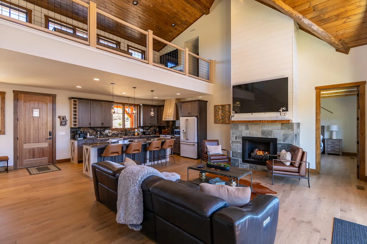 This image features an expansive great room with a stately stone fireplace as the centerpiece. Large windows offer breathtaking mountain views, while plush leather sofas invite relaxation. The open-concept layout seamlessly connects the living area with a modern, fully equipped kitchen and a spacious island with bar seating. Warm wooden beams and rustic accents complete the inviting lodge atmosphere.