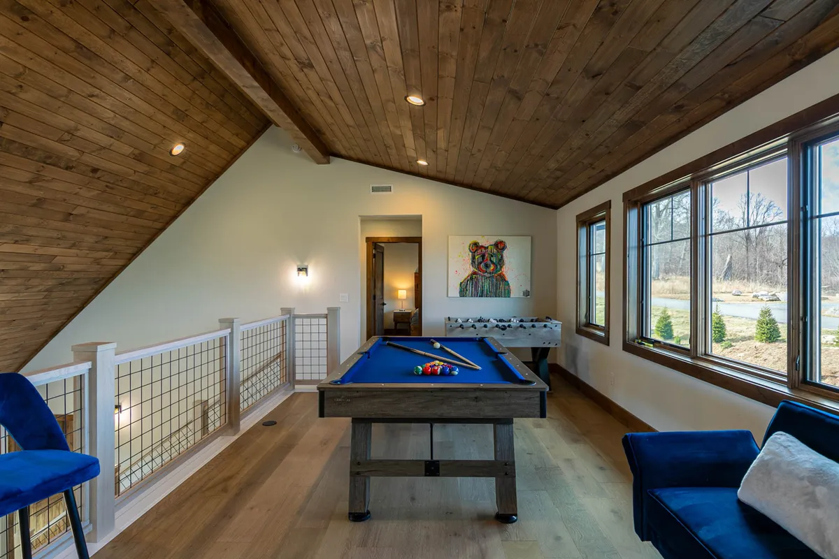 This image features a stylish game area with a pool table, a comfortable blue chair for spectators or reading, and striking artwork on the wall. Large windows fill the room with daylight, blending an energetic vibe with spaces for leisure and conversation.