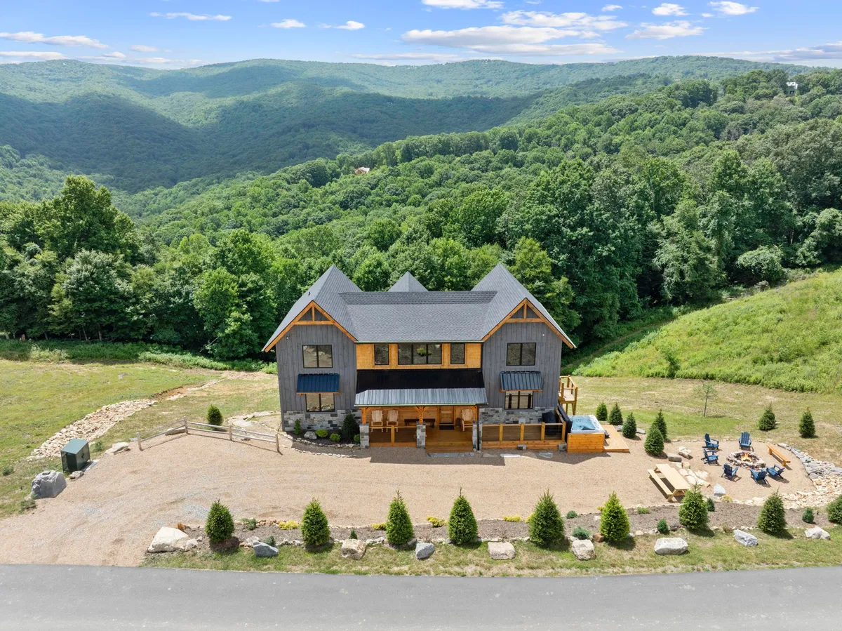 A striking modern cabin sits atop rolling green hills, surrounded by sweeping mountain vistas. The spacious outdoor seating area offers a serene spot to relax and take in the expansive views, while the gravel driveway welcomes guests to a luxurious mountain retreat.