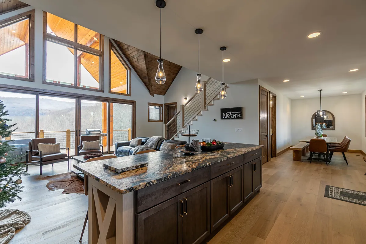 A welcoming space combining a comfortable seating area, a generous kitchen island, and a dining area all in one view. Large windows flood the room with natural light and offer panoramic mountain scenery. The arrangement encourages social interaction, cooking, and dining, reflecting the warmth and communal spirit of the lodge.